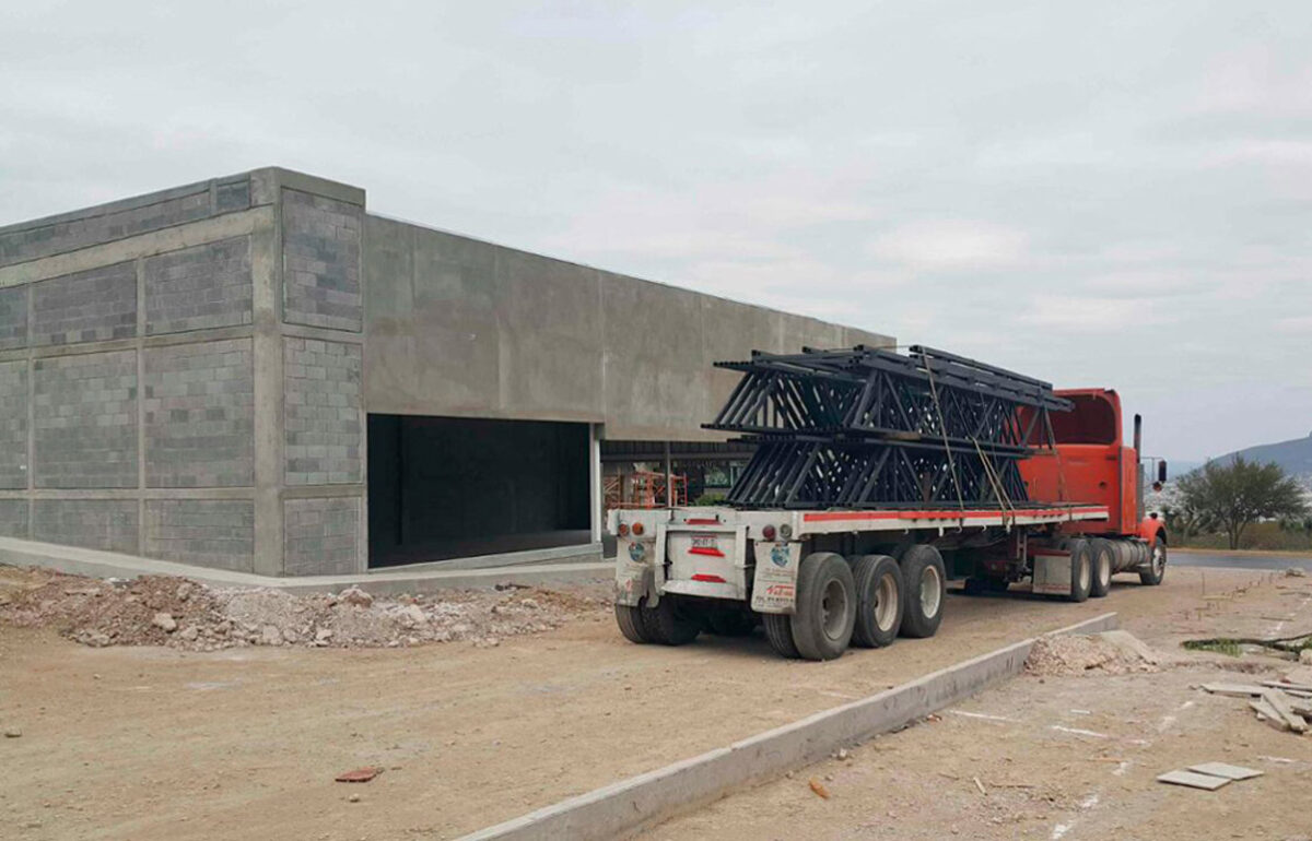 Tráiler con plataforma cargado de estructuras metálicas tipo joist en sitio de obra para construcción industrial.